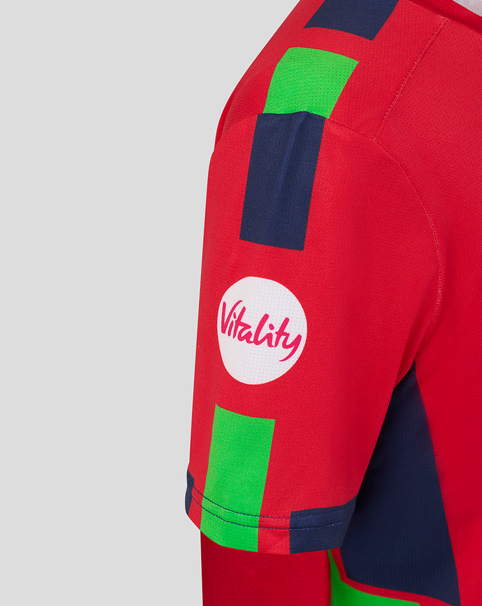 Side view of red junior T20 jersey showing navy and green graphic blocks and a white Vitality patch.